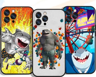 May include: Three phone cases with shark-themed designs. One case features a shark with a woman, another shows a shark with colorful decorations, and the third displays a shark in a suit with a Ferris wheel in the background.