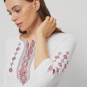 May include: White long-sleeved shirt with intricate red and black floral embroidery around the neckline and on the sleeves. The shirt features a round neckline with a drawstring. The sleeves are three-quarter length.