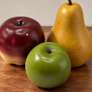 May include: A collection of three decorative fruit figurines including a dark red apple with a yellow base, a smooth green apple with the word JAPAN printed near the stem, and a textured yellow pear with a brown stem, all arranged together.