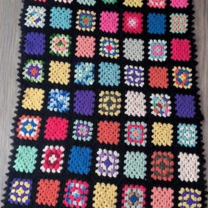 May include: A rectangular, handmade crochet piece featuring a patchwork design. The squares are in various colors, including pink, blue, yellow, and green, set against a black background. The edges have a scalloped border.