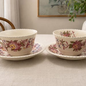 May include: Two cream-colored ceramic teacups and matching saucers featuring a scalloped edge and a detailed floral pattern in shades of pink, purple, and green. Each cup has a rounded handle and sits centered on its respective saucer.