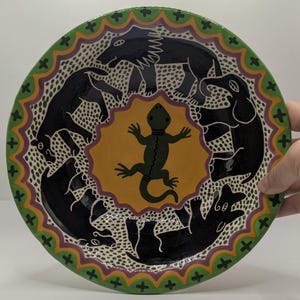 May include: A round ceramic plate featuring a central yellow star shape with a dark green lizard. Surrounding the center are four black animal figures, including a lion and an elephant, set against a white background with black dots. The rim is green.