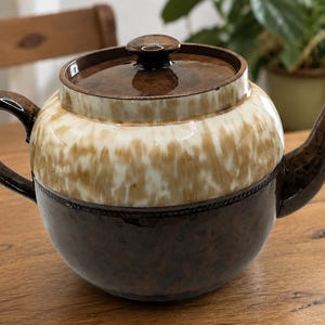 May include: A ceramic teapot featuring a two-tone glaze design. The upper portion displays a cream and tan drip pattern, while the lower section is a solid dark brown. It includes a matching lid with a knob, a curved handle, and a spout.