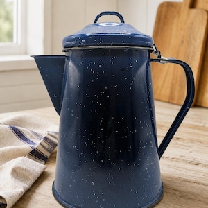 May include: A dark blue enamel coffee percolator featuring a white speckled pattern throughout the exterior. The vessel includes a matching lid with a small handle, a side handle, and a wide spout. The finish is smooth and glossy in appearance.