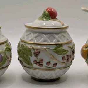 May include: Three white ceramic jars with lids and spoons. Each jar features a different fruit design: plums, berries, and oranges. The jars have gold trim and are decorated with embossed fruit and leaves.