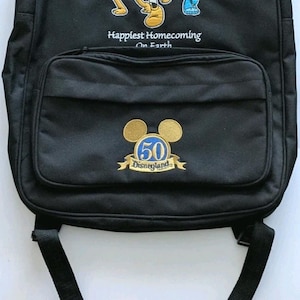 May include: Black backpack featuring a colorful embroidered design of Disney characters, including Mickey Mouse, Goofy, and Donald Duck. The backpack has a front pocket with a gold emblem that reads "50 Disneyland". The text "Happiest Homecoming On Earth" is also visible.