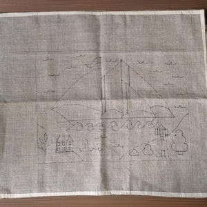 May include: A gray linen fabric with a hand-drawn sailboat scene. The design includes a sailboat, waves, a house, trees, and clouds. The edges of the fabric are bordered with a white trim. The artwork is done in black ink.