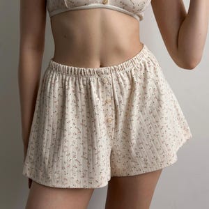 May include: Cream-colored pajama set with a floral pattern. The set includes a button-up bralette and matching shorts. The shorts have a button closure and an elastic waistband. The fabric has a ribbed texture.