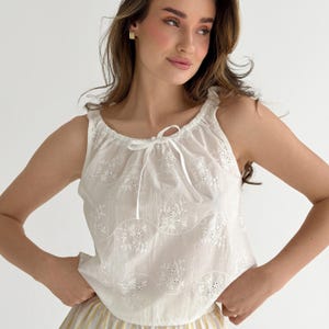 May include: A white sleeveless top featuring a round neckline with a drawstring tie closure. The fabric is adorned with horizontal wavy lines and delicate floral eyelet embroidery patterns throughout the bodice, creating a textured appearance.