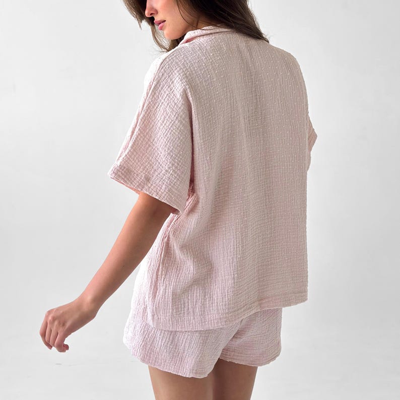 May include: A light pink pajama set, featuring a short-sleeved button-up shirt and matching shorts. The fabric has a textured, crinkled appearance. The set is designed for comfort and relaxation.