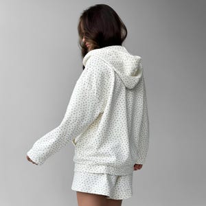 Op de afbeelding: Cr&egrave;mekleurige hoodie en short set met een delicaat, all-over bloemenpatroon in gedempt groen. De hoodie heeft een capuchon met trekkoord en een ritssluiting aan de voorkant. De shorts hebben een comfortabele, relaxte pasvorm.