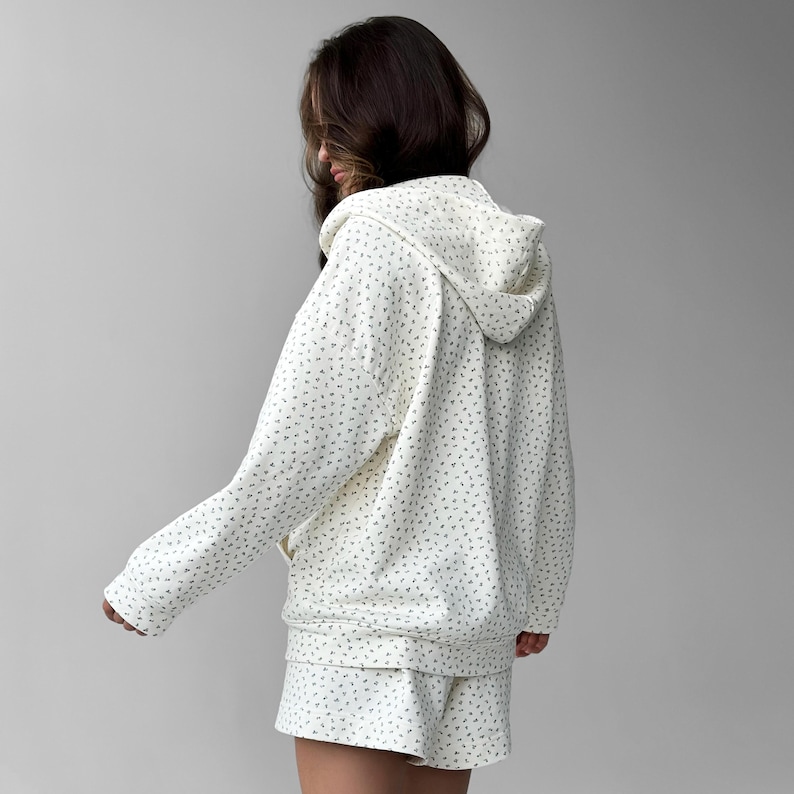 Op de afbeelding: Cr&egrave;mekleurige hoodie en short set met een delicaat, all-over bloemenpatroon in gedempt groen. De hoodie heeft een capuchon met trekkoord en een ritssluiting aan de voorkant. De shorts hebben een comfortabele, relaxte pasvorm.