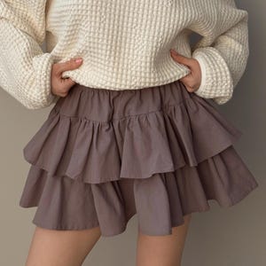 Peut inclure: Une jupe à volants de couleur mauve clair. La jupe est composée de trois niveaux de tissu, créant une silhouette volumineuse. Le modèle porte un pull en tricot gaufré crème. La jupe est une pièce élégante pour une tenue décontractée.