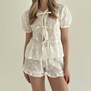 May include: White embroidered pajama set featuring a short-sleeved top with a tie closure and ruffled hem, paired with matching shorts. The set is adorned with a floral pattern and has a delicate, airy appearance. The fabric appears to be lightweight and semi-sheer.