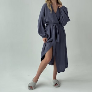 May include: A long, slate blue robe with a wrap-around design and a matching belt. The robe features long sleeves and a V-neckline. The model is wearing gray slippers.