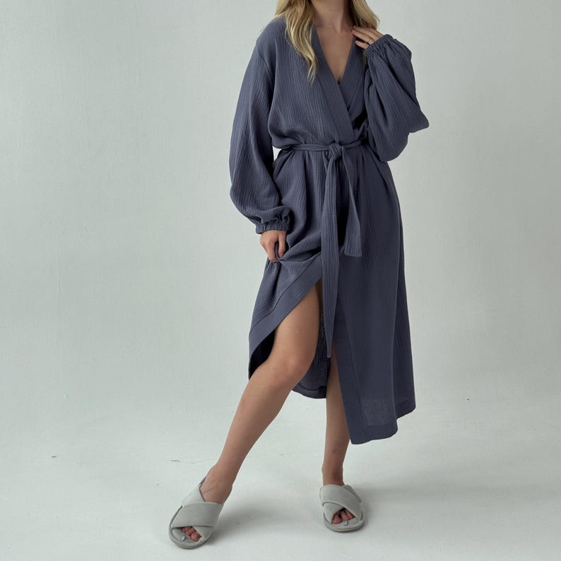 May include: A long, slate blue robe with a wrap-around design and a matching belt. The robe features long sleeves and a V-neckline. The model is wearing gray slippers.