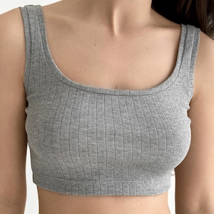 Könnte beinhalten: Graues geripptes Crop-Top mit rundem Ausschnitt und breiten Trägern. Das Oberteil hat ein strukturiertes, gemustertes Design und besteht aus einem weichen, dehnbaren Material. Das Kleidungsstück ist auf Komfort und Stil ausgelegt.