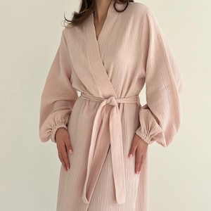 May include: A light pink robe with a wrap-around design and a matching belt tied at the waist. The robe features long sleeves with gathered cuffs. The fabric has a textured appearance.