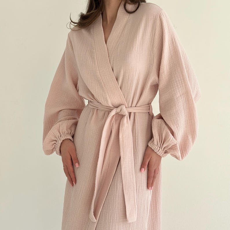 May include: A light pink robe with a wrap-around design and a matching belt tied at the waist. The robe features long sleeves with gathered cuffs. The fabric has a textured appearance.