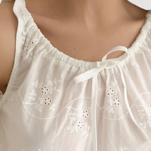 May include: A white sleeveless top featuring a gathered round neckline with a thin fabric tie closure. The garment is crafted from lightweight fabric adorned with intricate white floral eyelet embroidery and scalloped stitching details.
