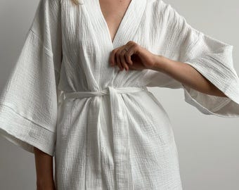 以下が含まれることがあります： 質感のあるシワ加工が施された白いローブ。幅広の七分袖と、ウエストで結ぶ共布のベルトが特徴です。Vネックラインとフロントのラップスタイルが、シンプルで清潔感のある外観を作り出しています。