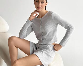May include: A light grey two-piece lounge set consisting of a long-sleeved button-down cardigan with a round neckline and matching high-waisted shorts. The fabric has a fine ribbed texture and a consistent heathered grey colour throughout the garment.