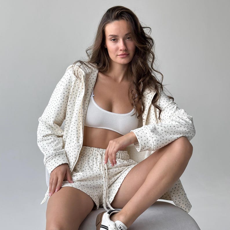 Op de afbeelding: Cr&egrave;mekleurige pyjamaset met een bloemenpatroon. De set bevat een jasje met ritssluiting, een short met trekkoord en een witte bh. Het model draagt witte sneakers met zwarte strepen.