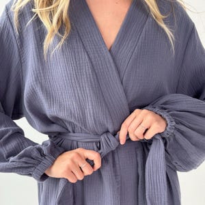 May include: A blue-grey robe with a wrap-around design and a matching belt. The robe features long sleeves with gathered cuffs. The fabric has a textured, ribbed appearance. The robe is being tied at the waist.