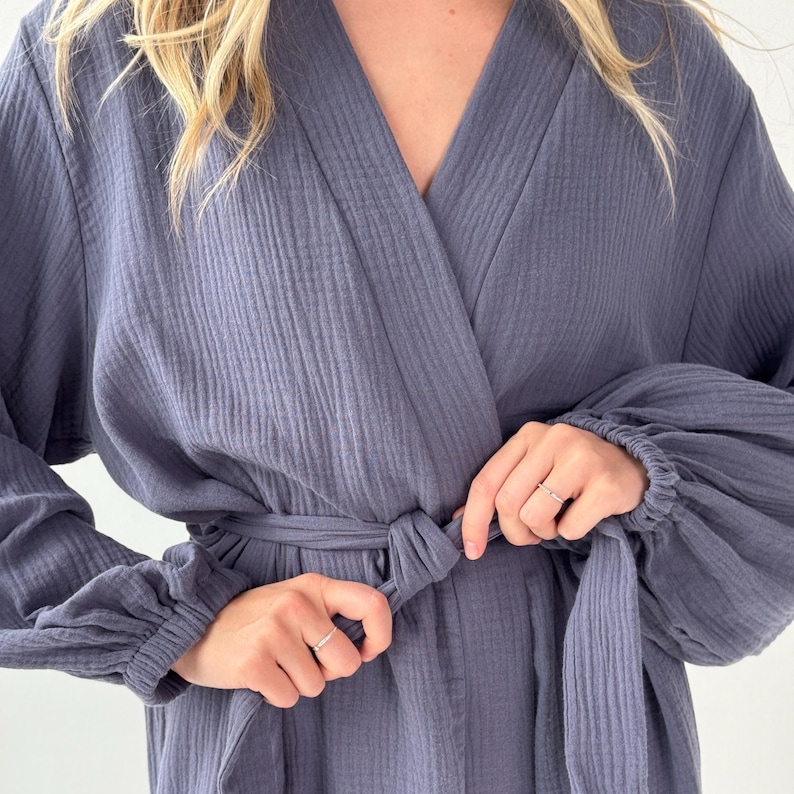 May include: A blue-grey robe with a wrap-around design and a matching belt. The robe features long sleeves with gathered cuffs. The fabric has a textured, ribbed appearance. The robe is being tied at the waist.