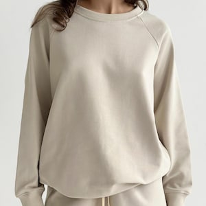 Könnte beinhalten: Ein beigefarbenes Sweatshirt mit Rundhalsausschnitt, langen Raglanärmeln, gerippten Bündchen und einem gerippten Halsausschnitt. Das Kleidungsstück besteht aus glattem, einfarbigem Stoff und wird mit einer passenden beigen Hose kombiniert.