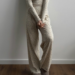 May include: Cream-colored floral print loungewear set. The long-sleeved top and wide-leg pants feature a delicate pattern of small, dark floral accents. The outfit is made of a soft, comfortable material, perfect for relaxing at home.