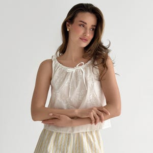 May include: A sleeveless white top featuring an eyelet lace floral pattern and a drawstring neckline with a thin white ribbon tie. The garment is paired with yellow and white vertically striped bottoms, creating a coordinated two-piece set.