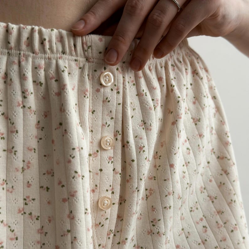 May include: Cream-colored, ribbed fabric with a delicate floral pattern in pink and green. The garment features a button-up design with three visible, round, cream-colored buttons. The elastic waistband is visible at the top.
