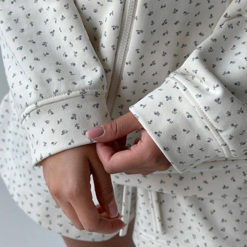 Op de afbeelding: Cr&egrave;mekleurige zip-up hoodie en bijpassende short set, versierd met een delicaat blauw bloemenpatroon. De manchetten en zoom zijn geribbeld. De outfit is gemaakt van een zacht, comfortabel materiaal, ideaal om te loungen of voor casual wear.