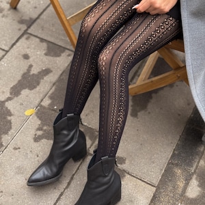 May include: Black patterned tights with a decorative design. The tights are paired with black leather ankle boots with a pointed toe and a small heel. The person is sitting on a wooden chair.