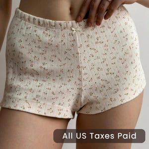 Op de afbeelding: Crèmekleurige shorts met een delicaat bloemenpatroon in roze en groen. De shorts hebben een geribbelde textuur en een klein strikdetail in de tailleband. De tekst "All US Taxes Paid" staat onderaan.
