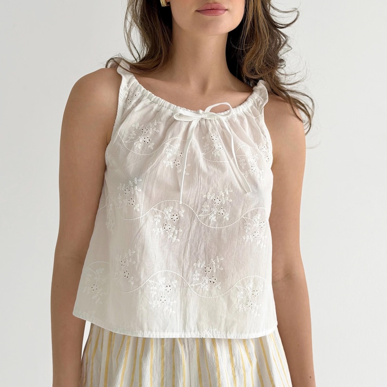 May include: A white sleeveless top featuring an eyelet lace floral pattern and a gathered neckline with a thin drawstring tie. The fabric displays horizontal wavy embroidered lines across the front, creating a textured and lightweight appearance.