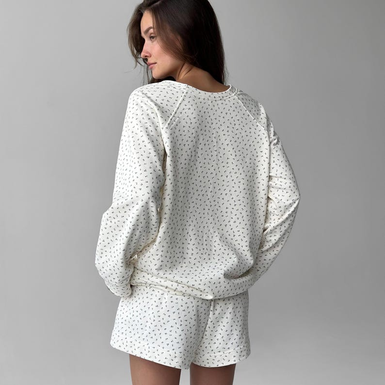 May include: Cream-colored pajama set with a repeating pattern of small, dark green shapes. The set includes a long-sleeved sweatshirt and matching shorts. The sweatshirt has a crew neck and the shorts have an elastic waistband.