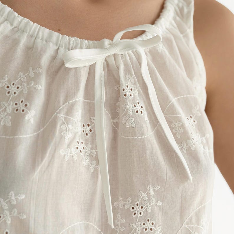 May include: A white sleeveless top featuring an eyelet embroidery pattern with floral motifs. The neckline is gathered with a drawstring closure tied into a bow at the center front, made from the same white fabric as the garment body.