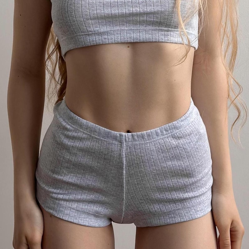 K&ouml;nnte beinhalten: Ein zweiteiliges Set aus hellgrauem Rippstrick, bestehend aus einem Crop-Top und passenden Shorts. Das Outfit ist aus weichem, dehnbarem Material gefertigt. Die Shorts sind hoch tailliert und haben eine bequeme Passform. Das Top ist schlicht und klassisch.