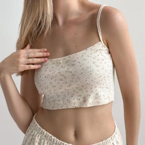 Op de afbeelding: Cr&egrave;mekleurige crop top met bloemenprint, dunne bandjes en een klein strikdetail. De top wordt gecombineerd met een bijpassende short, waardoor een geco&ouml;rdineerde set ontstaat. De stof heeft een geribbelde textuur en het bloemenpatroon bevat kleine roze en bruine accenten.