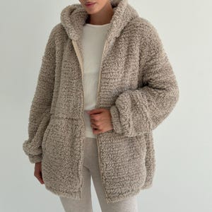 Peut inclure: Une veste à capuche zippée beige en matière polaire texturée et duveteuse. La veste présente des manches longues, une fermeture éclair intégrale sur le devant et une capuche. Elle est portée sur un haut blanc uni et un pantalon clair, douce au toucher.
