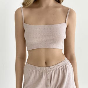 Op de afbeelding: Een lichtroze cropped hemdje met dunne spaghettibandjes en een vierkante halslijn, voorzien van een delicaat pointelle breipatroon. De top wordt gecombineerd met een bijpassende lichtroze short met een verticale ribbelstructuur en drie knopen.