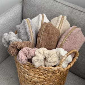 May include: A wicker basket containing several fluffy cosmetic bags and scrunchies. The bags are in shades of grey, brown, and pink, with gold-coloured zips. The scrunchies match the bags in colour and texture.