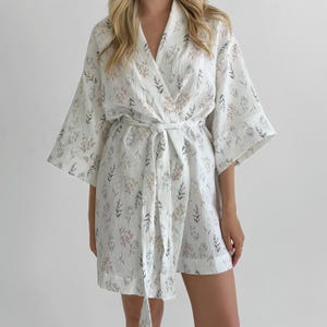 May include: A white waffle-textured robe featuring a delicate botanical print with small grey leaves and muted tan floral clusters. The garment has wide three-quarter length sleeves, a V-neckline, and a matching fabric tie belt at the waist.