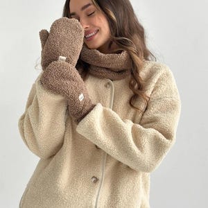 Könnte beinhalten: Ein Paar braune Sherpa-Fäustlinge mit einem kleinen weißen rechteckigen Etikett, ein passender brauner Sherpa-Halswärmer und eine cremefarbene Sherpa-Jacke mit Knöpfen. Die Jacke hat einen Rundhalsausschnitt, lange Ärmel und sichtbare Knöpfe.