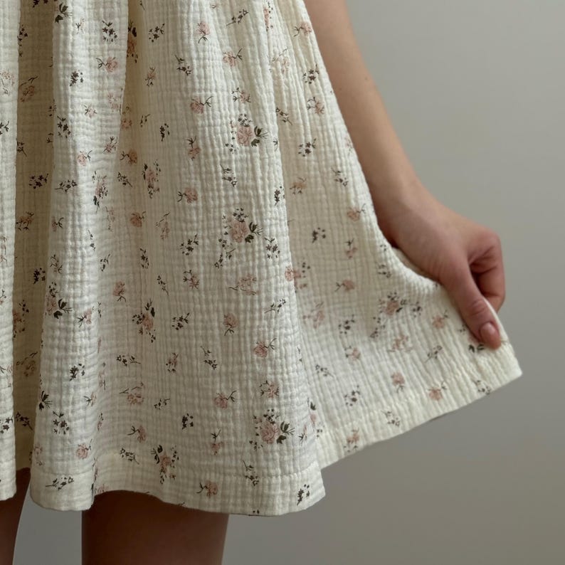 Op de afbeelding: Een cr&egrave;mekleurige jurk met een bloemenpatroon. De jurk heeft kleine roze bloemen en donkergroene bladeren verspreid over de stof. De stof heeft een getextureerde, licht gekreukelde uitstraling.