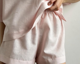 May include: A light pink linen shorts set. The shorts have an elasticated waistband and a relaxed fit. The top is a short-sleeved shirt with a collar. The fabric has a slightly textured appearance. The set is a solid colour.