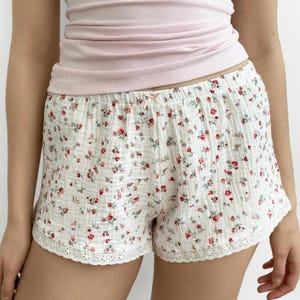 May include: White shorts featuring a small red and green floral pattern, a textured fabric finish, an elastic waistband with a small center bow, and a white eyelet lace trim along the bottom hem, paired with a light pink ribbed tank top.