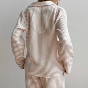 Op de afbeelding: Een beige en wit gestreepte pyjamaset. De top met lange mouwen heeft een kraag en een knoopsluiting. De bijpassende broek is ook gestreept. De stof lijkt een zacht, lichtgewicht materiaal te zijn.